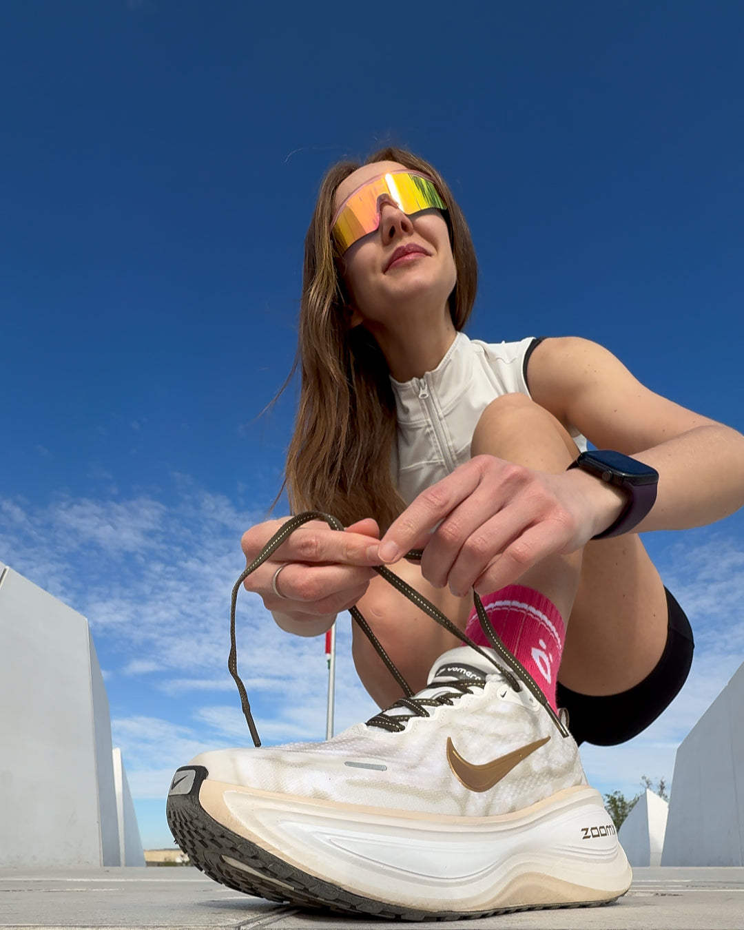 A woman wearing iridescent höhen aero sports sunglasses and pink höhen merino wool socks core while tying her running shoes outdoors under a blue sky
