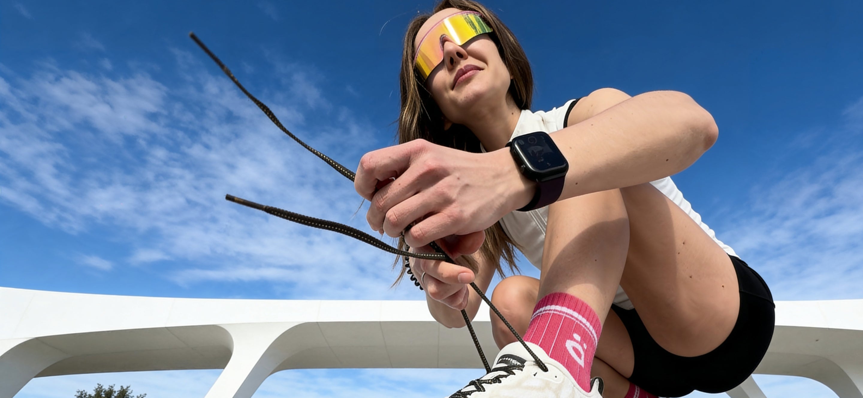 Woman outdoors wearing höhen Aero women sports sunglasses with mirrored lenses and pink Core Merino socks while tying running shoes under a bright blue sky.