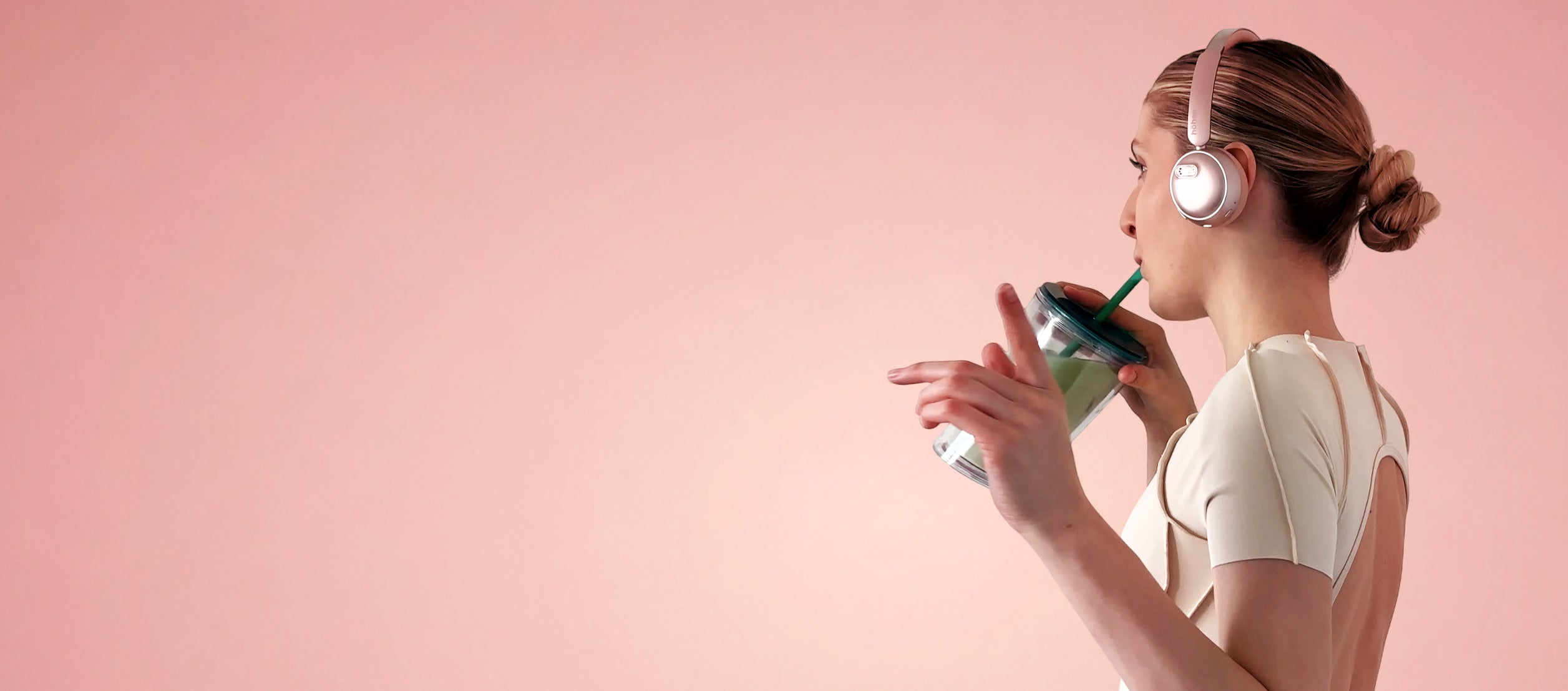 Side view desktop banner of a woman wearing rose gold höhen aura mini wireless headphones and drinking a green smoothie against a solid pink background with generous copy space on the left