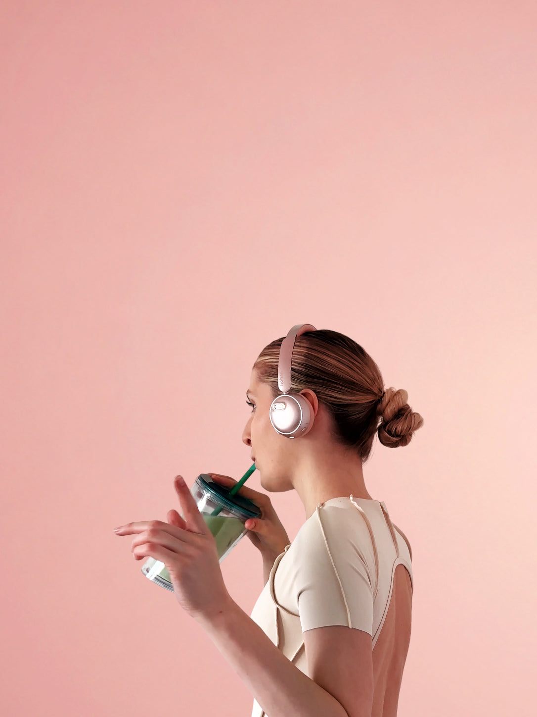 Vertical mobile banner of a woman in profile wearing rose gold höhen aura mini wireless headphones while holding a green drink against a clean pink background