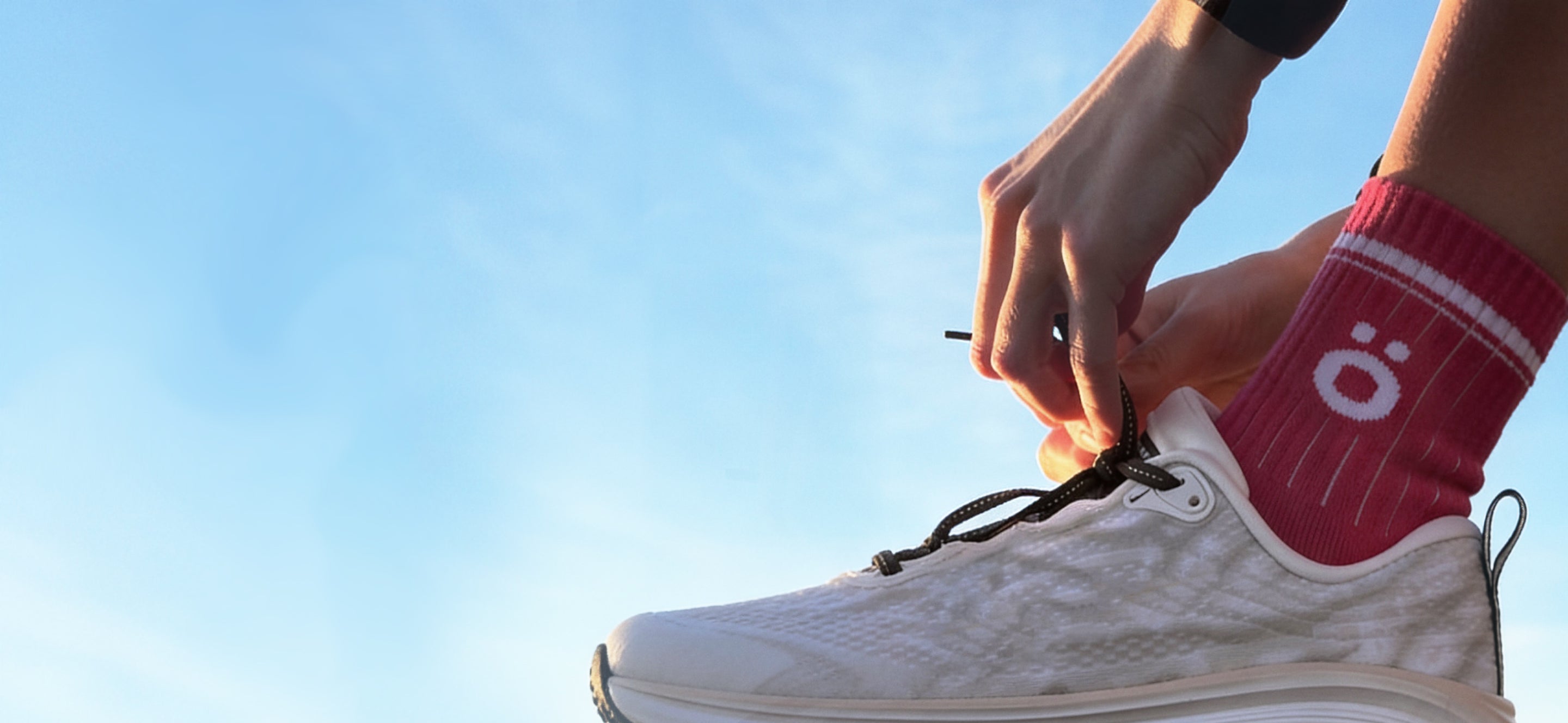 Close-up of a person tying white running shoes while wearing magenta höhen Core Merino socks outdoors under a clear blue sky