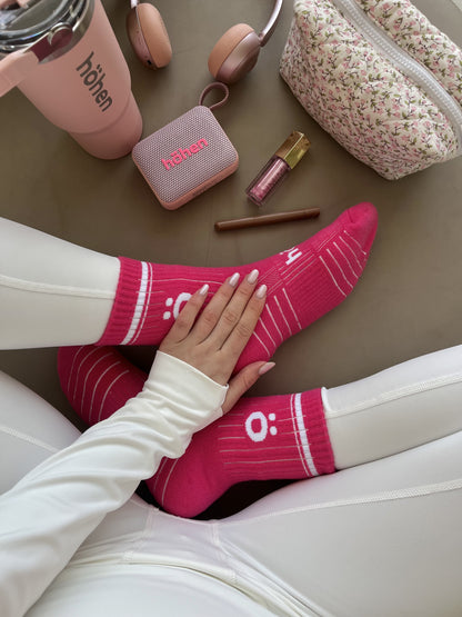 Top-down lifestyle view of a woman wearing magenta höhen core athletic socks, surrounded by a matching pink höhen essential tumbler, höhen move wireless portable speaker, and höhen aura mini headphones.