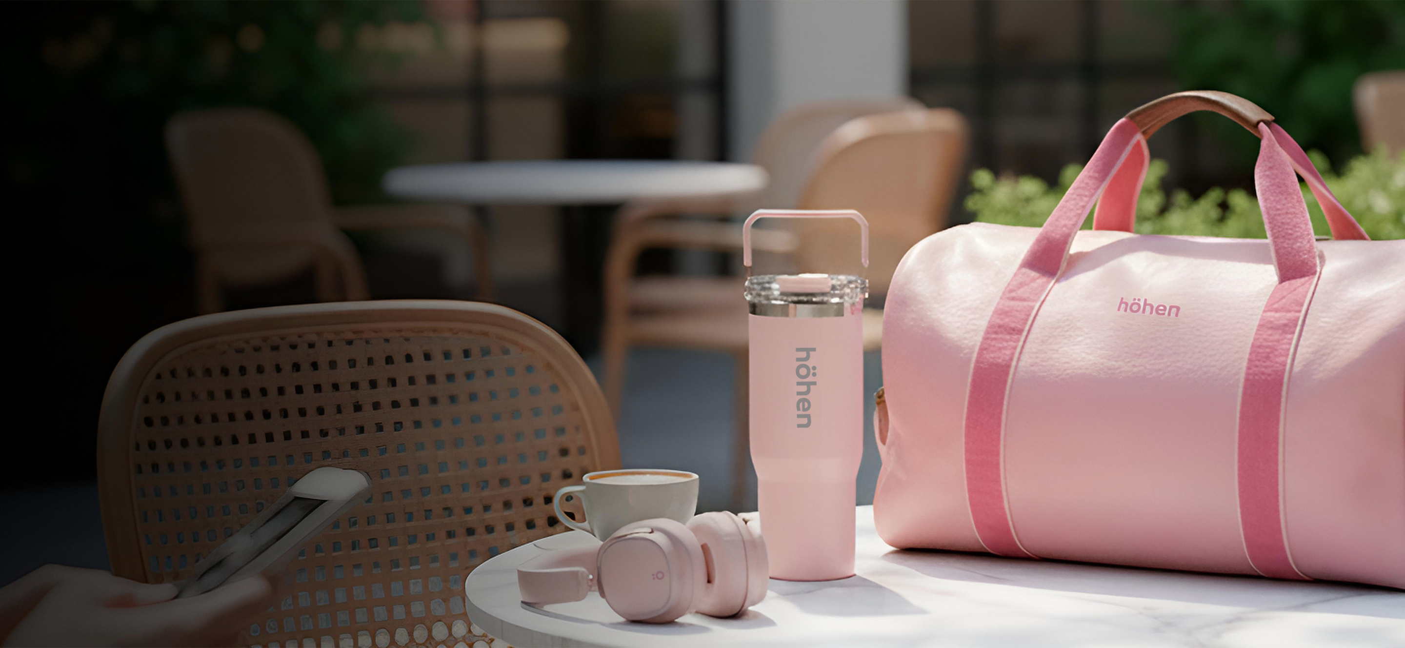 A collection of höhen products on a table: Aura Pro headphones, Essential Tumbler, and One bag, all in pastel pink.