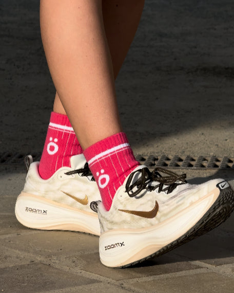Close-up of magenta 
höhen merino wool socks core with white logo worn with white and gold running shoes on a paved path