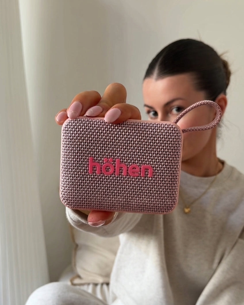 A close-up shot of a model holding the pink höhen Move portable speaker, showcasing its woven technical texture and prominent brand logo in a soft lifestyle setting