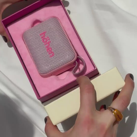 A woman unboxes the pink höhen move speaker from its premium pink packaging, then holds it poolside alongside the matching pink höhen essential tumbler and wireless headphones.