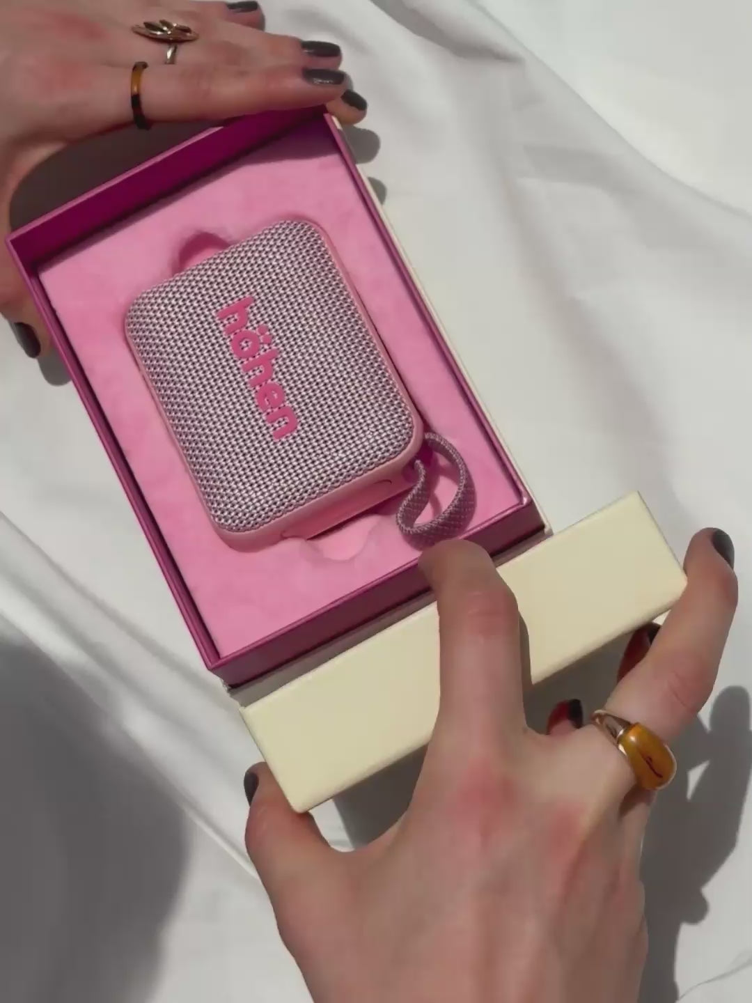A woman unboxes the pink höhen move speaker from its premium pink packaging, then holds it poolside alongside the matching pink höhen essential tumbler and wireless headphones.
